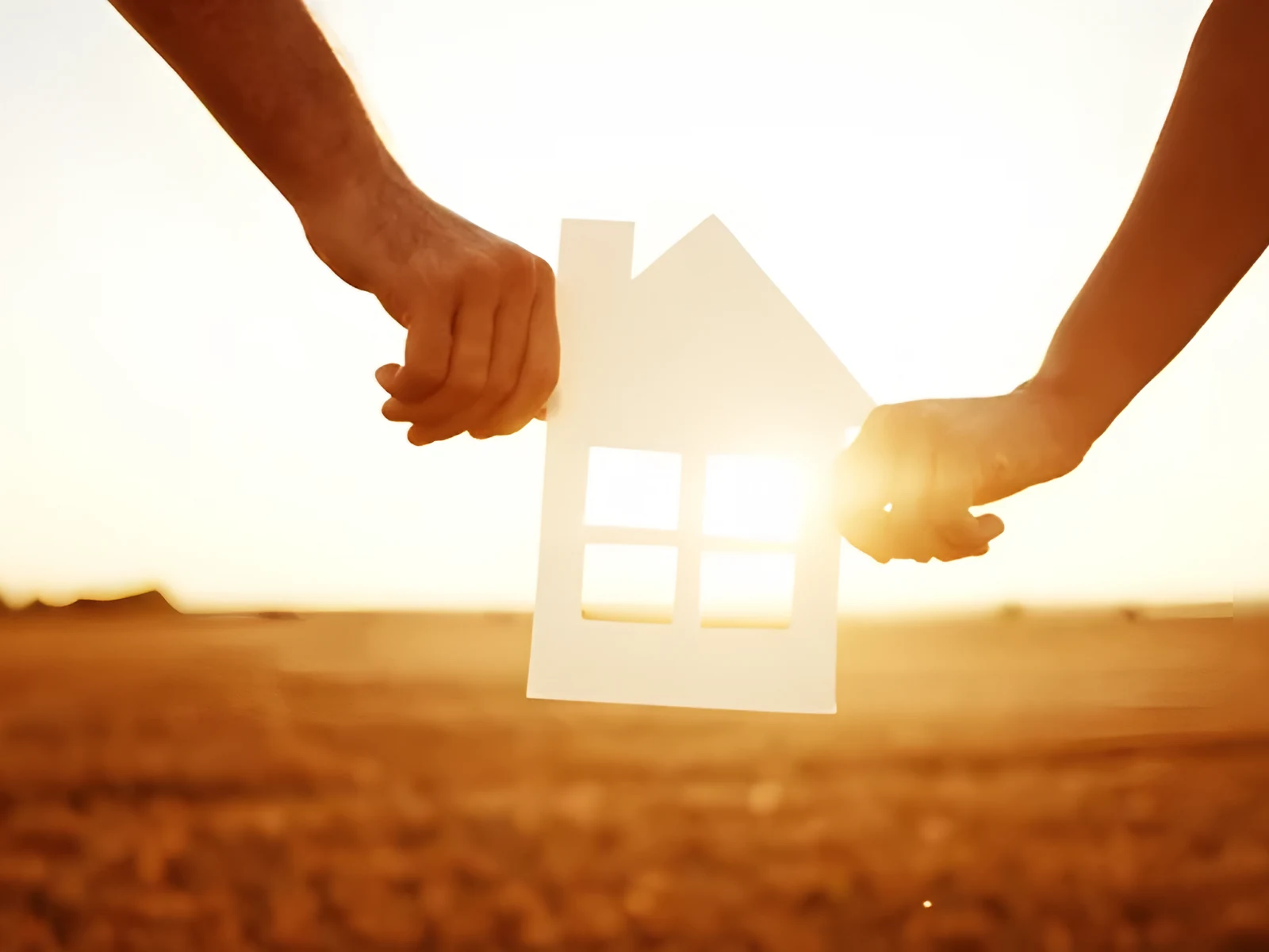 Zwei Hände halten ein Haus aus Papier vor der Abendsonne